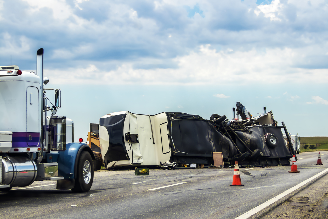 Fifth wheel RV overturned on highway with wench truck trying to get it off the road and two semis parked nearby and traffic cones keeping traffic away